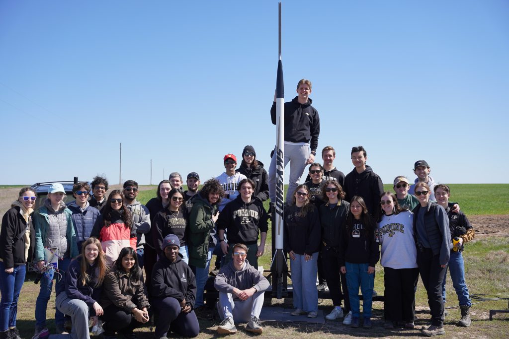 HA Skyshot team photo Students pose for a photo in a field around a 8 foot tall rocket that has "skyshot" written on the side