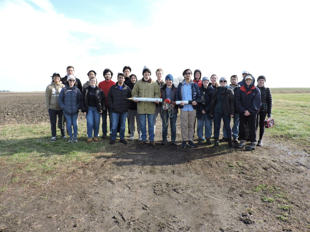 SL holding rocket A group of students stand in a muddy field holding a white rocket horizontally