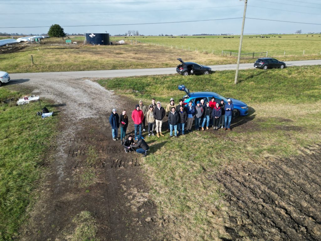 Spaceport IRI launch drone Aerial picture of a group of students standing in a muddy field next to a car