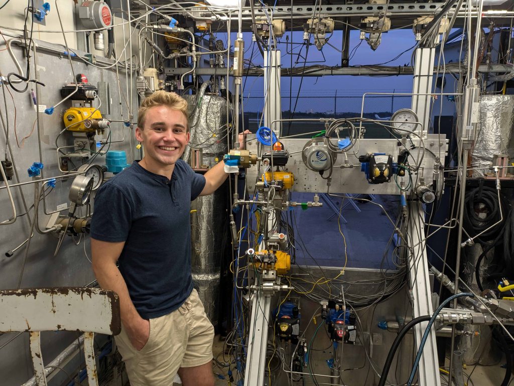 LQ S25 newsletter 4 A student with blond hair stands amidst a lot of fluid systems covering the floor and walls